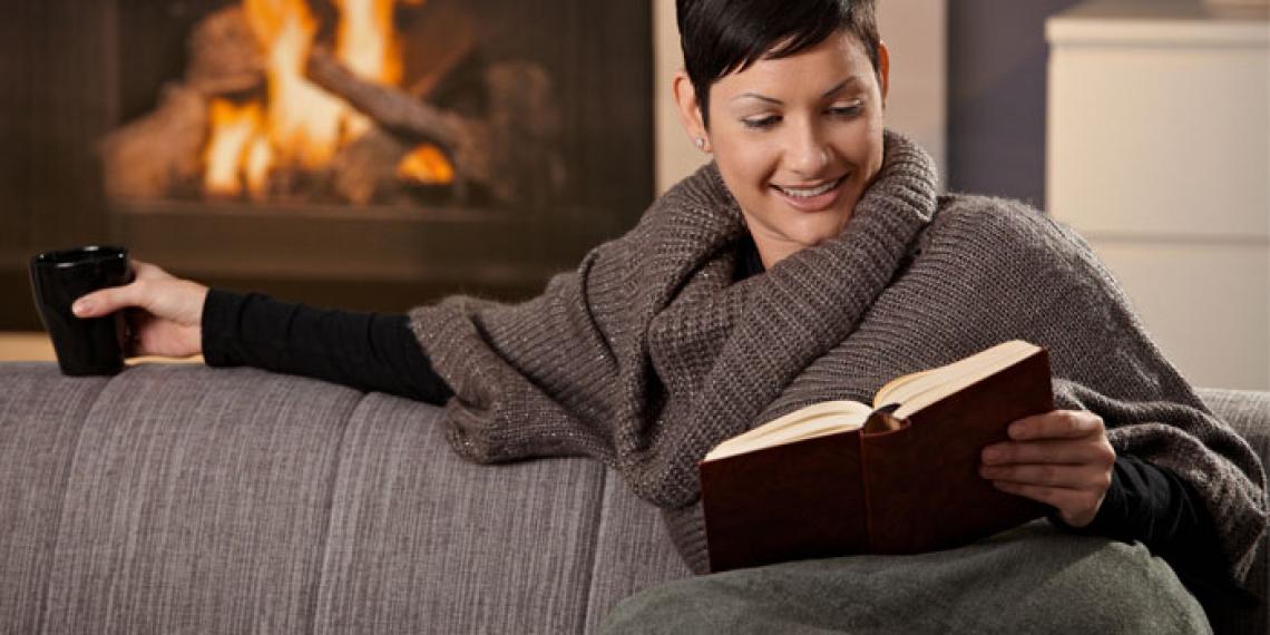 a woman in a blanket reading a book by a fire